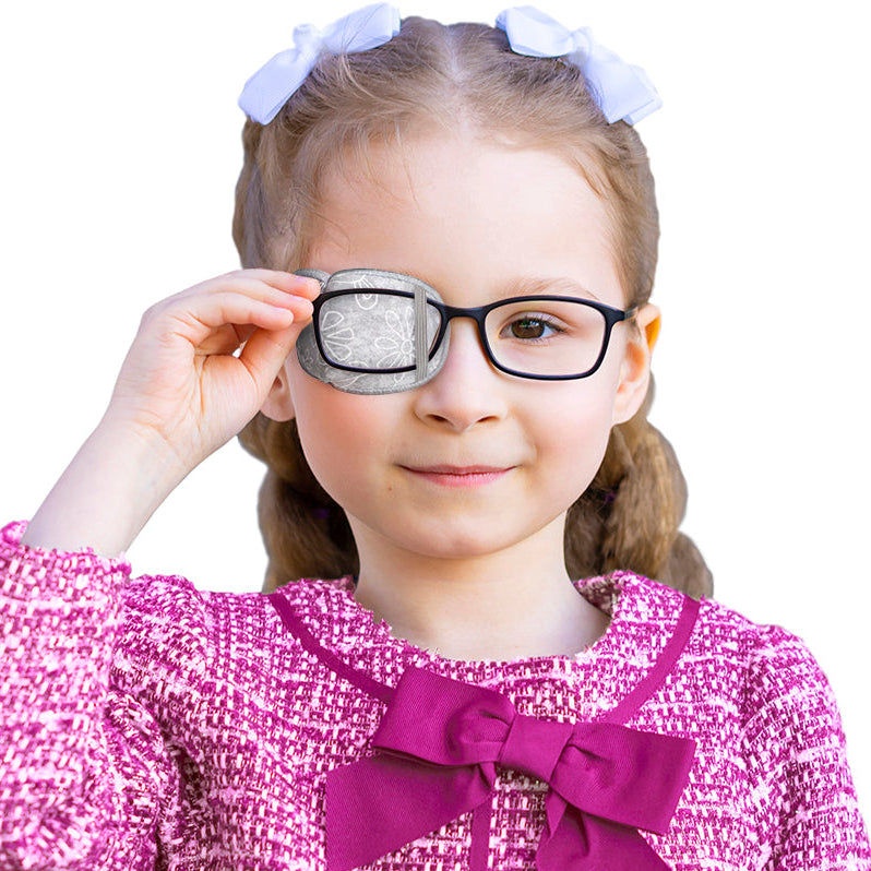 A young girl with light brown hair in pigtails and white bows, wearing a pink tweed top with a purple bow, holding one of her black-rimmed glasses to her eye against a plain white background.