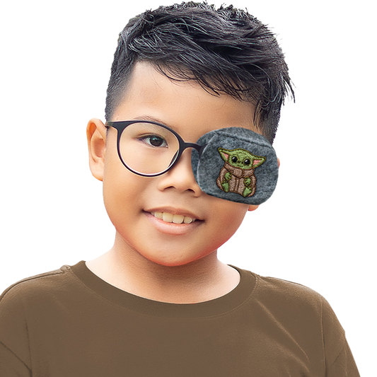 A young boy wearing a brown long-sleeve shirt and round black eyeglasses, with an eye patch over one eye featuring a green embroidered character on a gray background, set against a solid white studio backdrop.